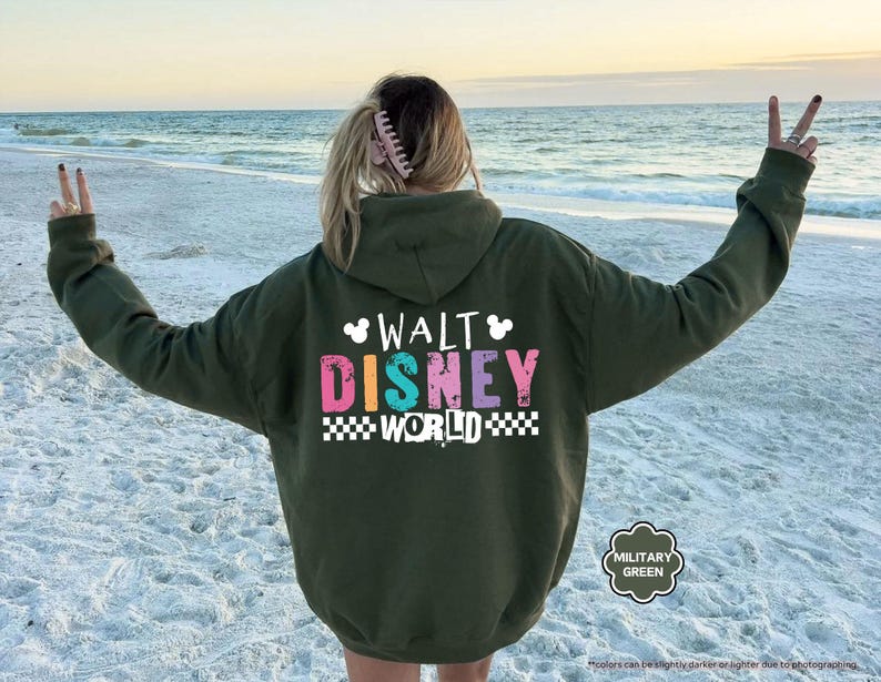 Puede incluir: Sudadera con capucha verde militar con el texto "WALT DISNEY WORLD" en letras de bloque coloridas. La sudadera tiene una capucha con cord&oacute;n y un bolsillo de canguro. El fondo es una playa con el oc&eacute;ano.