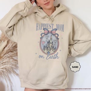 May include: Sand-colored hoodie with a front graphic. The graphic features a castle design within a decorative oval, with the text "HAPPIEST MOM on Earth" above. A light pink bow and Mickey Mouse heads are also included in the design.