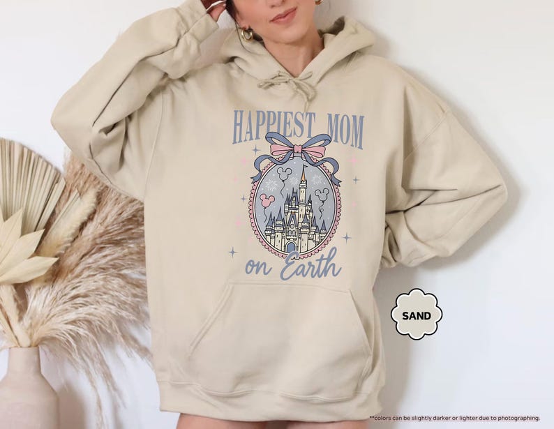 May include: Sand-colored hoodie with a front graphic. The graphic features a castle design within a decorative oval, with the text "HAPPIEST MOM on Earth" above. A light pink bow and Mickey Mouse heads are also included in the design.