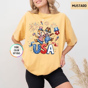 May include: Mustard-colored t-shirt featuring a cartoon graphic of two chipmunks celebrating with fireworks and the word "USA" in red, white, and blue. The shirt has a relaxed fit and short sleeves. The words "More Color Options" are also visible.