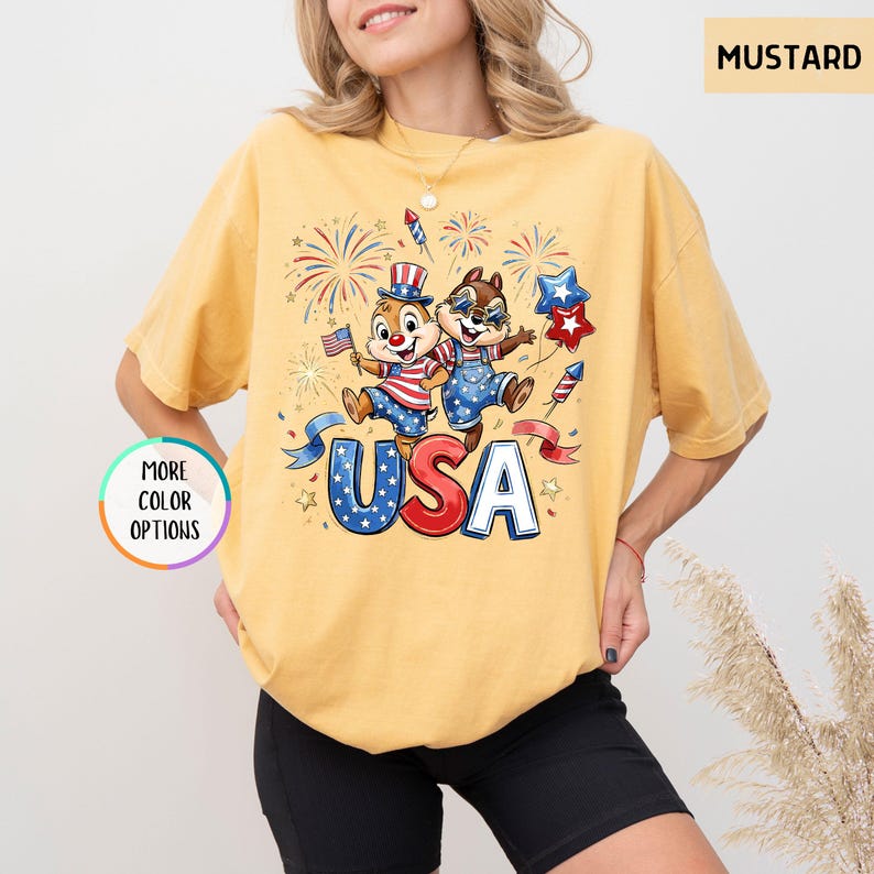 May include: Mustard-colored t-shirt featuring a cartoon graphic of two chipmunks celebrating with fireworks and the word "USA" in red, white, and blue. The shirt has a relaxed fit and short sleeves. The words "More Color Options" are also visible.