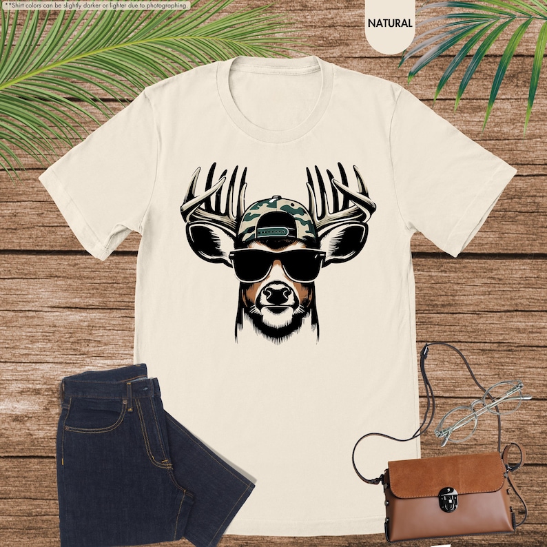 May include: A white t-shirt with a graphic of a deer wearing a camouflage baseball cap and sunglasses. The text "NATURAL" is printed in the top right corner of the image.