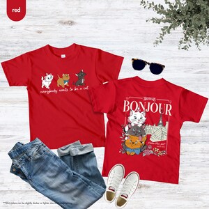 May include: Two red t-shirts with cat graphics. One shirt says "everybody wants to be a cat" with cartoon cats. The other shirt says "Bonjour" with a graphic of Marie and other cats. Includes blue jeans, white sneakers, and sunglasses.