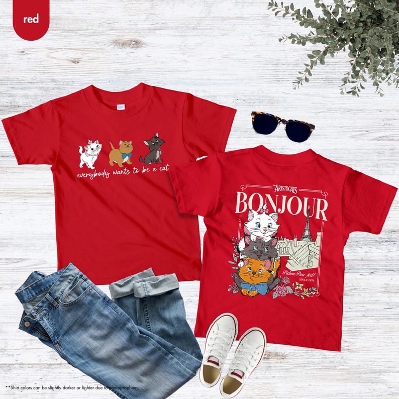May include: Two red t-shirts with cat graphics. One shirt says "everybody wants to be a cat" with cartoon cats. The other shirt says "Bonjour" with a graphic of Marie and other cats. Includes blue jeans, white sneakers, and sunglasses.
