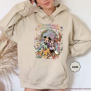 May include: Sand-colored hoodie featuring a colorful graphic of Disney characters, including Mickey and Minnie Mouse, surrounded by flowers and the text "EPCOT International Flower & Garden Festival". The hoodie has a front pocket and a hood.