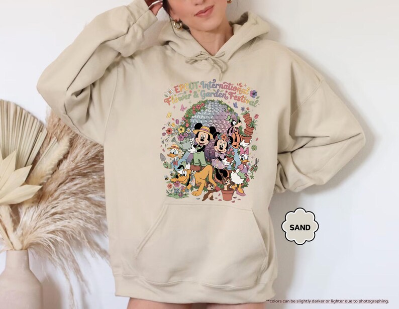 May include: Sand-colored hoodie featuring a colorful graphic of Disney characters, including Mickey and Minnie Mouse, surrounded by flowers and the text "EPCOT International Flower & Garden Festival". The hoodie has a front pocket and a hood.