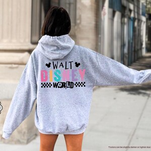 Puede incluir: Una sudadera con capucha gris claro con el texto "WALT DISNEY WORLD" en letras coloridas y un patr&oacute;n de cuadros blanco y negro. La sudadera con capucha tiene una capucha y mangas largas. Las palabras "SPORT GREY" est&aacute;n en un c&iacute;rculo gris.