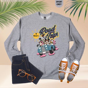 Op de afbeelding: Sportgrijs sweatshirt met een Disney-afbeelding van personages in een blauwe auto met de tekst "Road Trip! Miami". De afbeelding bevat een donkere spijkerbroek, een oranje bril en oranje sneakers.
