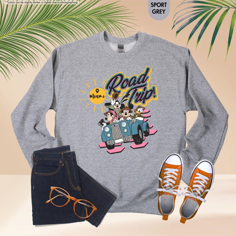 Op de afbeelding: Sportgrijs sweatshirt met een Disney-afbeelding van personages in een blauwe auto met de tekst "Road Trip! Miami". De afbeelding bevat een donkere spijkerbroek, een oranje bril en oranje sneakers.