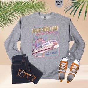 May include: Sport grey sweatshirt with a graphic that reads "PLEASE STAND CLEAR of the Doors" with a monorail and fireworks design. The sweatshirt is paired with dark wash jeans and orange sneakers.