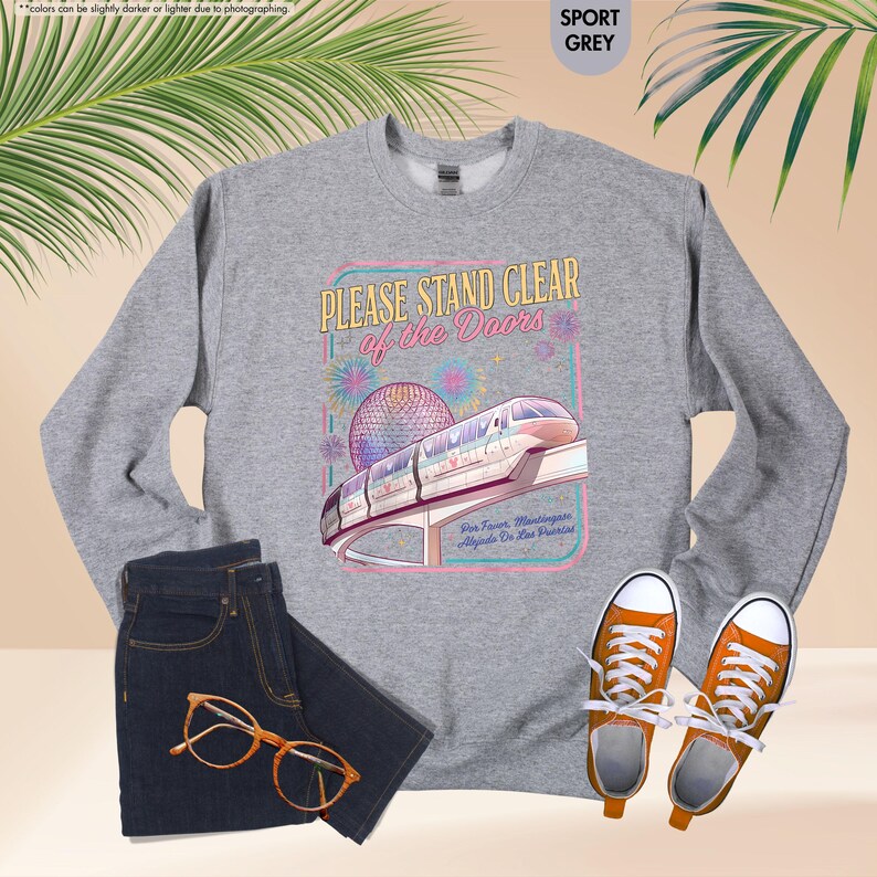 May include: Sport grey sweatshirt with a graphic that reads "PLEASE STAND CLEAR of the Doors" with a monorail and fireworks design. The sweatshirt is paired with dark wash jeans and orange sneakers.