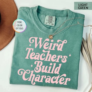May include: Light green t-shirt with the phrase "Weird Teachers Build Character" in pink, retro-style lettering. A gold necklace with a pendant is draped on the shirt. The shirt is a Comfort Colors brand, size S-CH.