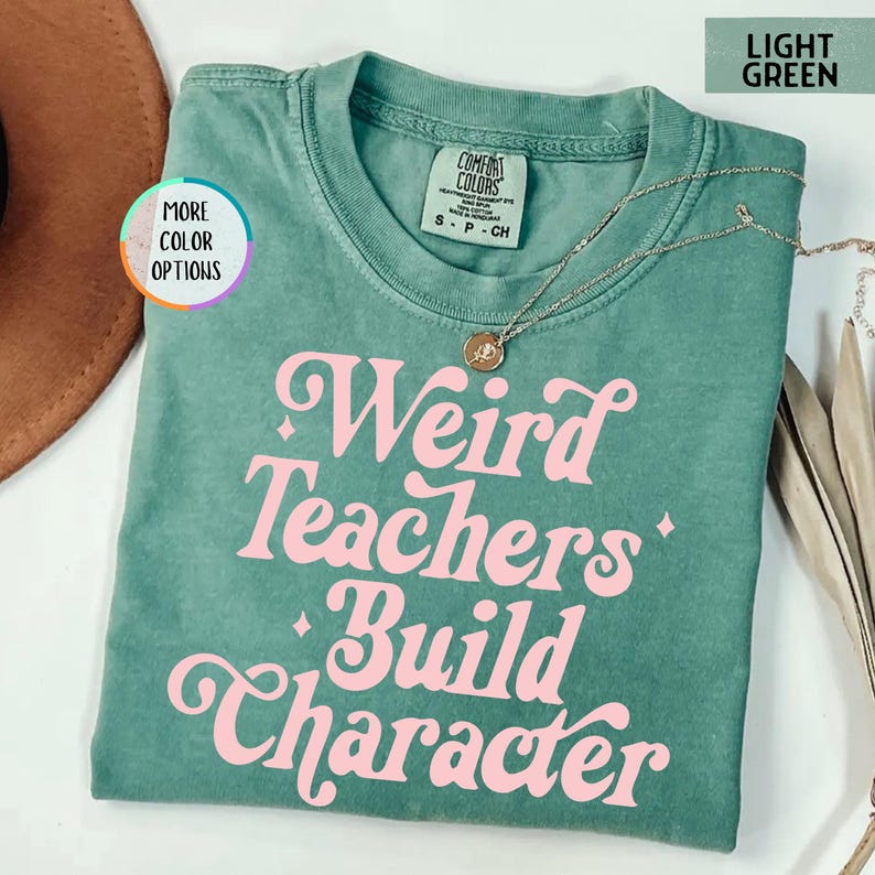 May include: Light green t-shirt with the phrase "Weird Teachers Build Character" in pink, retro-style lettering. A gold necklace with a pendant is draped on the shirt. The shirt is a Comfort Colors brand, size S-CH.