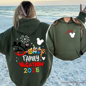 K&ouml;nnte beinhalten: Gr&uuml;ner Kapuzenpullover mit einem Disney Family Vacation 2025-Motiv auf der R&uuml;ckseite und einem Mickey Mouse-Motiv auf der Vorderseite. Der Pullover hat eine Kapuze mit Kordelzug und eine K&auml;ngurutasche.