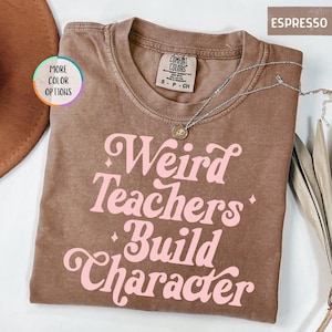 May include: Espresso-colored t-shirt with the phrase "Weird Teachers Build Character" in pink, retro-style lettering. The shirt has a crew neck and short sleeves. A silver necklace with a pendant is draped across the shirt.