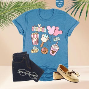 Puede incluir: Una camiseta azul con un gr&aacute;fico de art&iacute;culos con tem&aacute;tica de Mickey Mouse, que incluyen palomitas de ma&iacute;z, un helado de Mickey Mouse, globos y un boleto que dice "Magic". El texto "Magical" est&aacute; impreso en una pancarta en la parte superior del gr&aacute;fico.