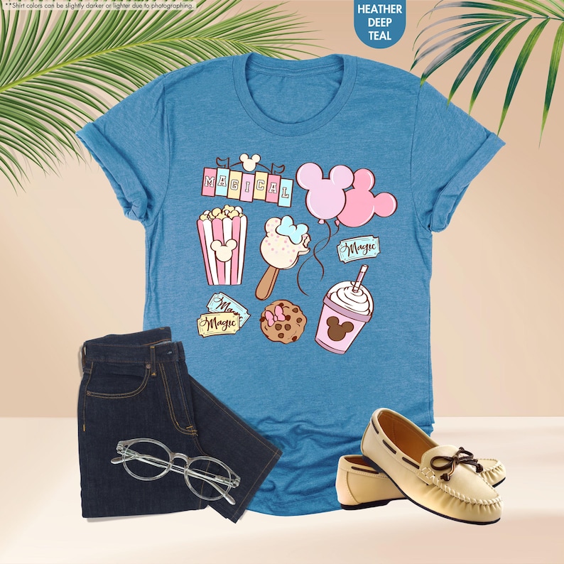 Puede incluir: Una camiseta azul con un gr&aacute;fico de art&iacute;culos con tem&aacute;tica de Mickey Mouse, que incluyen palomitas de ma&iacute;z, un helado de Mickey Mouse, globos y un boleto que dice "Magic". El texto "Magical" est&aacute; impreso en una pancarta en la parte superior del gr&aacute;fico.