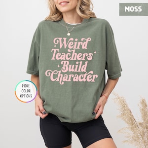 May include: Moss green t-shirt with the text "Weird Teachers Build Character" in pink script. The shirt is a relaxed fit and has short sleeves. The model is wearing black shorts and a necklace.