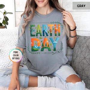 May include: A gray t-shirt with a colorful graphic design that says "Earth Day". The graphic features a stylized depiction of a forest, mountains, and clouds.
