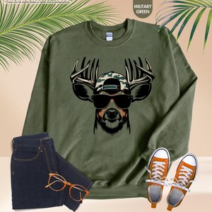May include: A green sweatshirt with a graphic of a deer wearing sunglasses and a camouflage hat. The deer has large antlers.