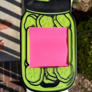 Può includere: Un supporto per foglietti adesivi a forma di barattolo di cetriolini stampato in 3D, color verde lime con coperchio e contorni neri. Il barattolo presenta un intaglio quadrato centrale per un post-it rosa acceso e dettagli di cetriolini in rilievo.