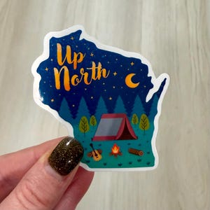 May include: A sticker in the shape of Wisconsin with a camping scene. The design features a dark blue night sky with the words "Up North" in orange, a tent, trees, a campfire, a guitar, and a crescent moon.