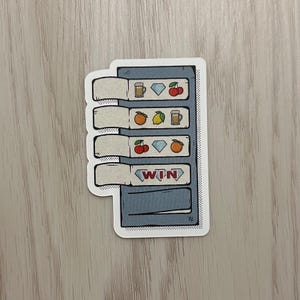 May include: A sticker featuring a cartoon slot machine design. The machine has four reels with symbols like cherries, diamonds, oranges, lemons, and beer mugs. The bottom reel displays the word "WIN" in a red banner.
