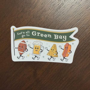 May include: A rectangular sticker featuring a green banner with the text Let's all go to Green Bay. Below the banner are four cartoon characters with arms and legs: a brown football, a beer mug, a yellow wedge of cheese, and a hot dog.