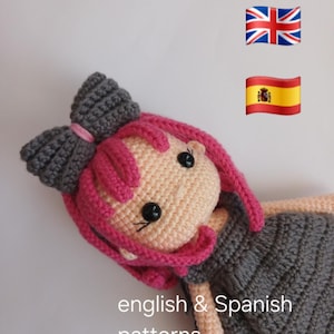 Könnte beinhalten: Handgefertigte Häkelpuppe mit rosa Haaren und einer grauen Schleife. Die Puppe hat schwarze Knopfaugen und trägt ein graues Kleid. Der Text "english & Spanish patterns" ist sichtbar.