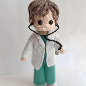 Könnte beinhalten: Handgemachte Häkelpuppe, die als Arzt gekleidet ist und einen weißen Kittel, türkisfarbene Arbeitskleidung und graue Schuhe trägt. Die Puppe hat braunes Haar und ein Stethoskop um den Hals. Die Puppe ist etwa 20 cm groß.