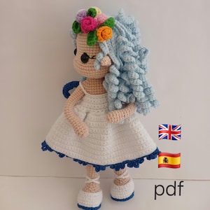 May include: A handmade crochet doll with blue curly hair, wearing a white dress with blue trim and ballet flats. The doll has a floral crown with pink, orange, and purple flowers. The doll's eyes are black, and the text "pdf" is visible.