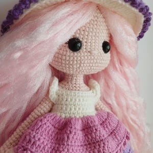 Op de afbeelding: Een gehaakte pop met roze haar, een witte en paarse hoed en een roze en witte jurk.