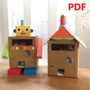 May include: Two cardboard boxes decorated as a robot and a house. The robot has a yellow head, green arms, and a brown body. The house has a red roof and a green door. Both boxes have clear plastic windows and a small hole in the bottom.