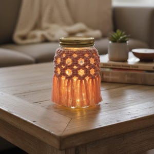 May include: A decorative jar with a woven, peach-colored macrame sleeve and a gold lid. The jar is lit from within, creating a warm glow. The jar sits on a wooden table, suggesting a cozy home decor item.