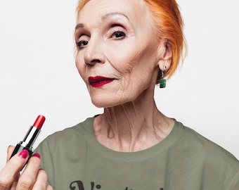 May include: An older person with orange hair and red lipstick holds a tube of red lipstick. They wear a green t-shirt with the word "Vintage" printed on it. The person is also wearing gold rings and green earrings.