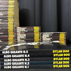 Può includere: Una collezione di fumetti di Dylan Dog, con la serie "Albo Gigante". Le copertine sono nere con testo bianco, che mostra titoli e numeri di uscita. Pile di altri numeri sono visibili sullo sfondo, creando un'esposizione completa della serie a fumetti.