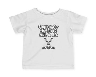 May include: White baby t-shirt with black text that reads "Eligible for the 2042 NHL Draft" and a graphic of two crossed hockey sticks and a puck.