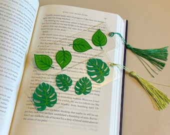 May include: Clear acrylic leaf-shaped bookmarks with green leaf designs and green tassels. The bookmarks are placed on the pages of a book. The text "WISH YOU WERE HERE" is visible on the book's page.