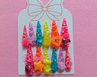 May include: A set of twelve long, pointed press-on nails in various bright colors, each adorned with decorative elements like rhinestones, bows, and flowers. The nails are arranged on a white card with a bow-shaped cutout, set against a pink background.