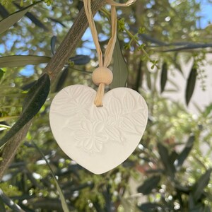 Peut inclure: Ornement en forme de cœur en céramique blanche avec un motif floral suspendu à une branche avec une ficelle en ficelle brune et une perle en bois.