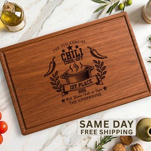 May include: A rectangular wooden cutting board featuring a laser-engraved design that reads THE 5TH ANNUAL CHILI COOK-OFF 1ST PLACE 2026 BEST DAMN CHILI IN TOWN THE ANDERSONS, accompanied by illustrations of a chili pot, peppers, and stars.
