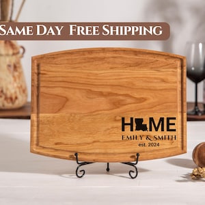 May include: A rectangular wooden cutting board with a juice groove and a black metal stand. The word "HOME" is printed on the board, with the Louisiana state outline replacing the "O". The text "EMILY & SMITH est. 2024" is below.