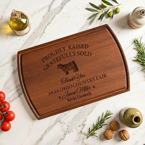 May include: A rectangular wooden cutting board with rounded corners and a juice groove, engraved with the text PROUDLY RAISED GRATEFULLY SOLD, a cow silhouette, Thank You, 2026 OHIO COUNTRY FAIR, Jamie Miller, and Kevin Livestock.