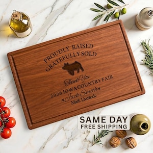 May include: A rectangular wooden cutting board with a juice groove, engraved with the text PROUDLY RAISED GRATEFULLY SOLD, a pig silhouette, Thank You, 2026 IOWA COUNTRY FAIR, Jack Smith, and Mark Livestock, surrounded by olive branches and tomatoes.