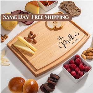 May include: A personalized wooden cutting board with the text "the Millers Est. 2024" engraved on it. The board is light brown and has a rounded edge. It is surrounded by various food items, including cheese, almonds, raspberries, and dried fruit.
