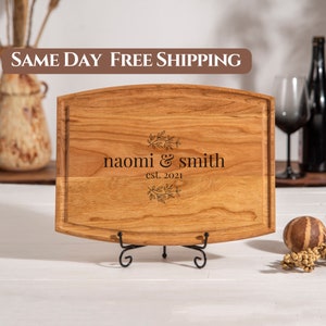 May include: A personalized wooden cutting board with a curved edge and a black metal stand. The board is engraved with the text "naomi & smith est. 2021" and a floral design.