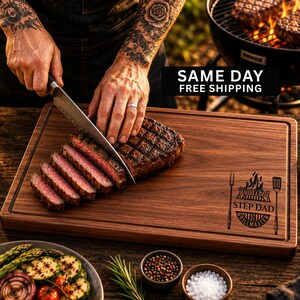 May include: A rectangular dark brown wooden cutting board with a juice groove and a laser-engraved design featuring a grill, a fork, a spatula, and the text "World's best STEP DAD GRILLMASTER". The board is used to slice a grilled steak.