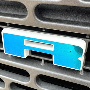 Peut inclure: Gros plan d'un emblème de voiture avec la lettre "R" blanche et bleue. La lettre est placée sur un fond de grille grise. La section bleue de la lettre a une qualité réfléchissante et irisée.