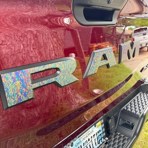 Op de afbeelding: Close-up van een bordeauxrode truckbed met het woord "RAM" in grote, iriserende letters. De letters hebben een textuur, kleurrijk patroon. Een zwarte handgreep en een kentekenplaat zijn ook zichtbaar.