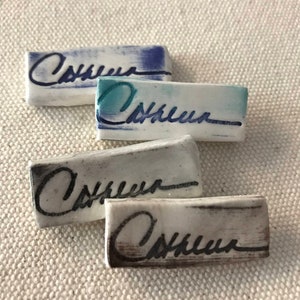May include: Four rectangular ceramic tiles with the name "Cathleen" written in cursive. The tiles are white with blue, teal, gray, and brown accents. The background is a textured, off-white fabric.